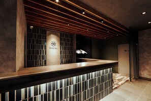 A hotel reception area with a wooden reception desk, a logo on the wall, and a patterned floor. - R Hotel Namba Daikokucho (Osaka)