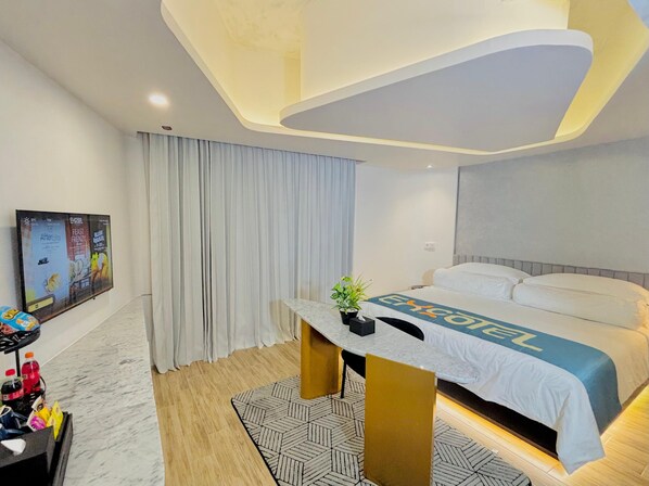 A modern hotel room with a large bed, a desk with a plant, and a wall-mounted TV displaying event posters. - Excotel Design Hotel  (Surabaya)