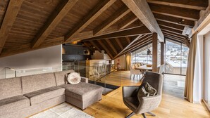 A spacious loft with a high ceiling, wooden beams, a modern sofa, a cushioned armchair, and a view of a snowy landscape through large windows.