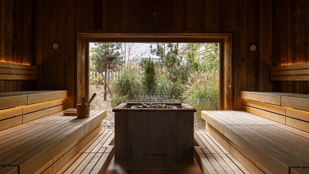 A sauna with wooden benches and a central heating element, surrounded by wooden walls and a view of trees.