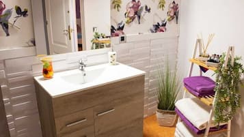 Un baño con un lavamanos blanco, un mueble de madera y un espejo. Se aprecia un papel tapiz floral, una planta y una repisa con botellas.