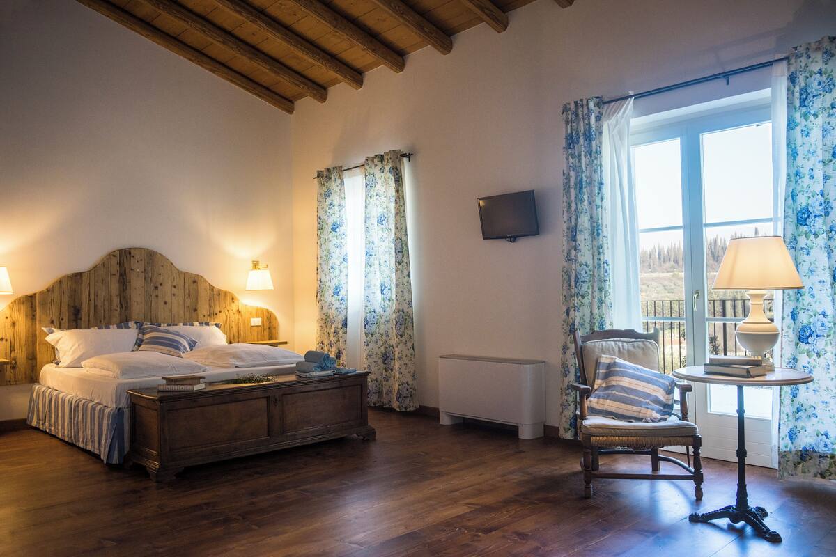 Ein Schlafzimmer mit Bettgestell aus Holz, einem Nachttisch mit Lampe, einem Sessel mit Kissen und einem Fenster mit floralem Vorhang.