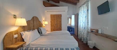 Une chambre à coucher avec une tête de lit en bois, deux lits avec une literie à rayures, une étagère en bois, un téléviseur et une salle de bain visible à travers une porte ouverte.