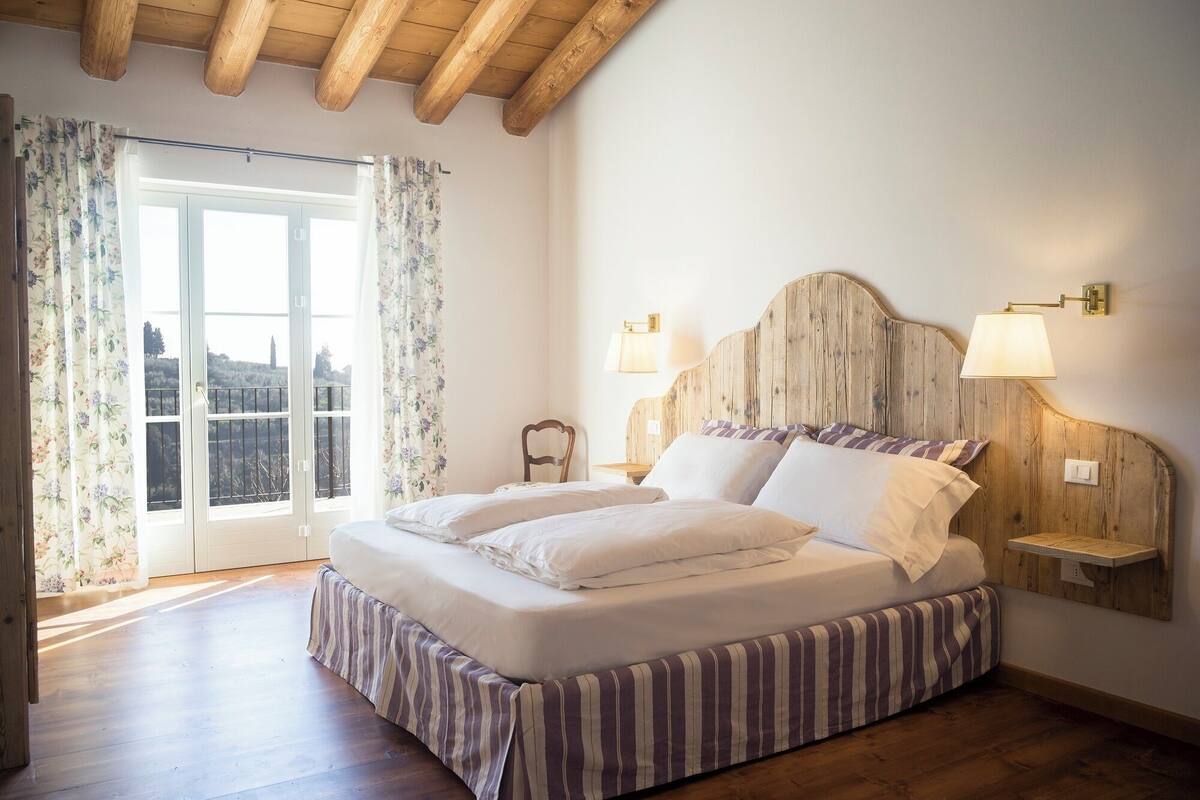 Ein Schlafzimmer mit einem großen Bett, einem hölzernen Kopfteil, zwei Nachttischlampen und Blick durch eine Glastür.