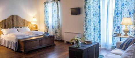 Quarto com cama de cabeceira de madeira, mesa de cabeceira com abajur, pequena mesa de cabeceira com vaso, sofá e radiador.