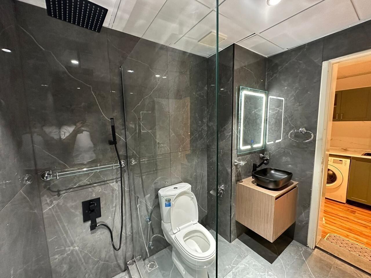 Un baño moderno con ducha envuelta en vidrio, inodoro blanco y tocador de madera con lavamanos negro.