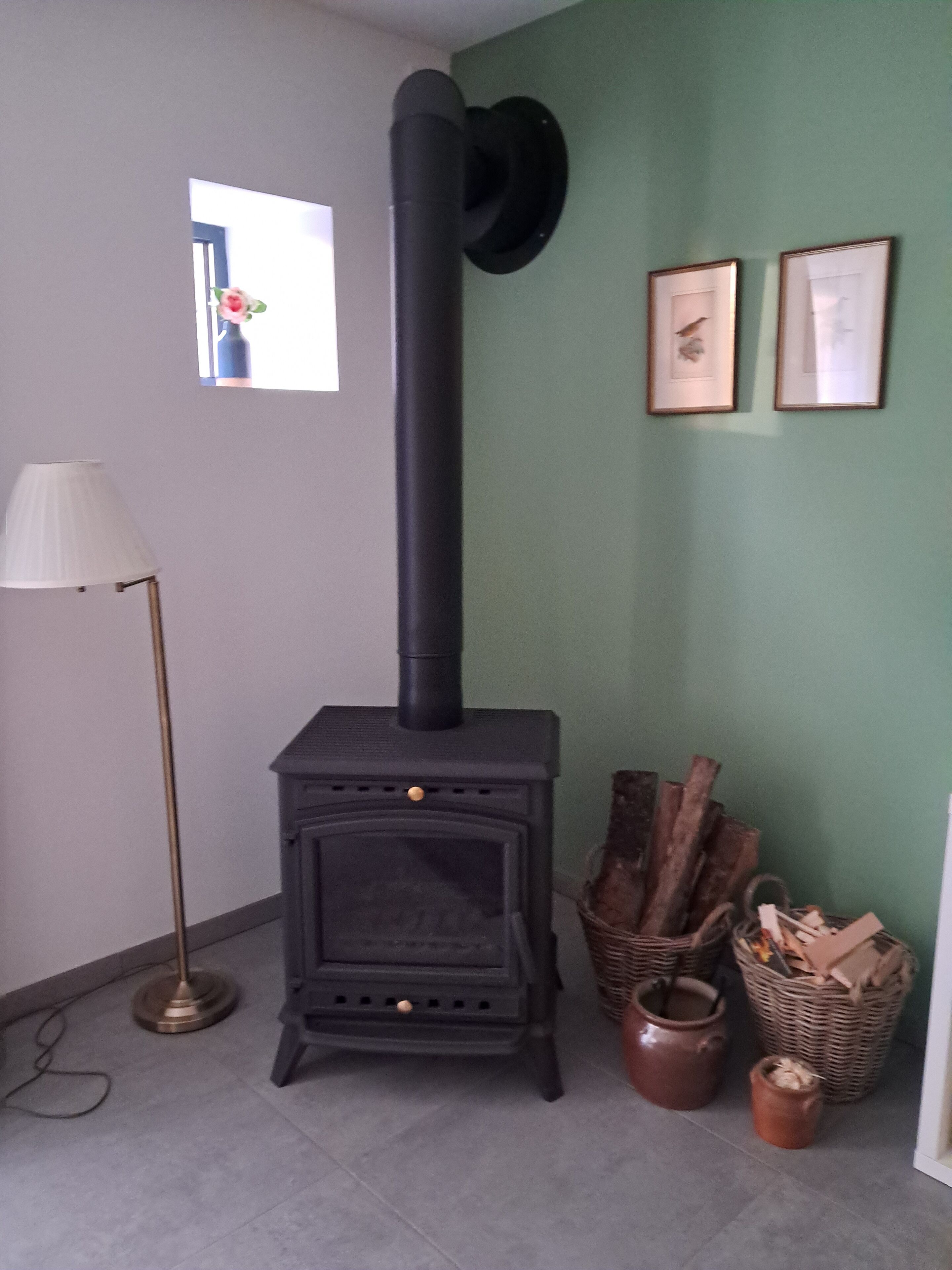 Una habitación con un estufa de leña negra, una lámpara de pie y una pared verde con dos cuadros enmarcados.