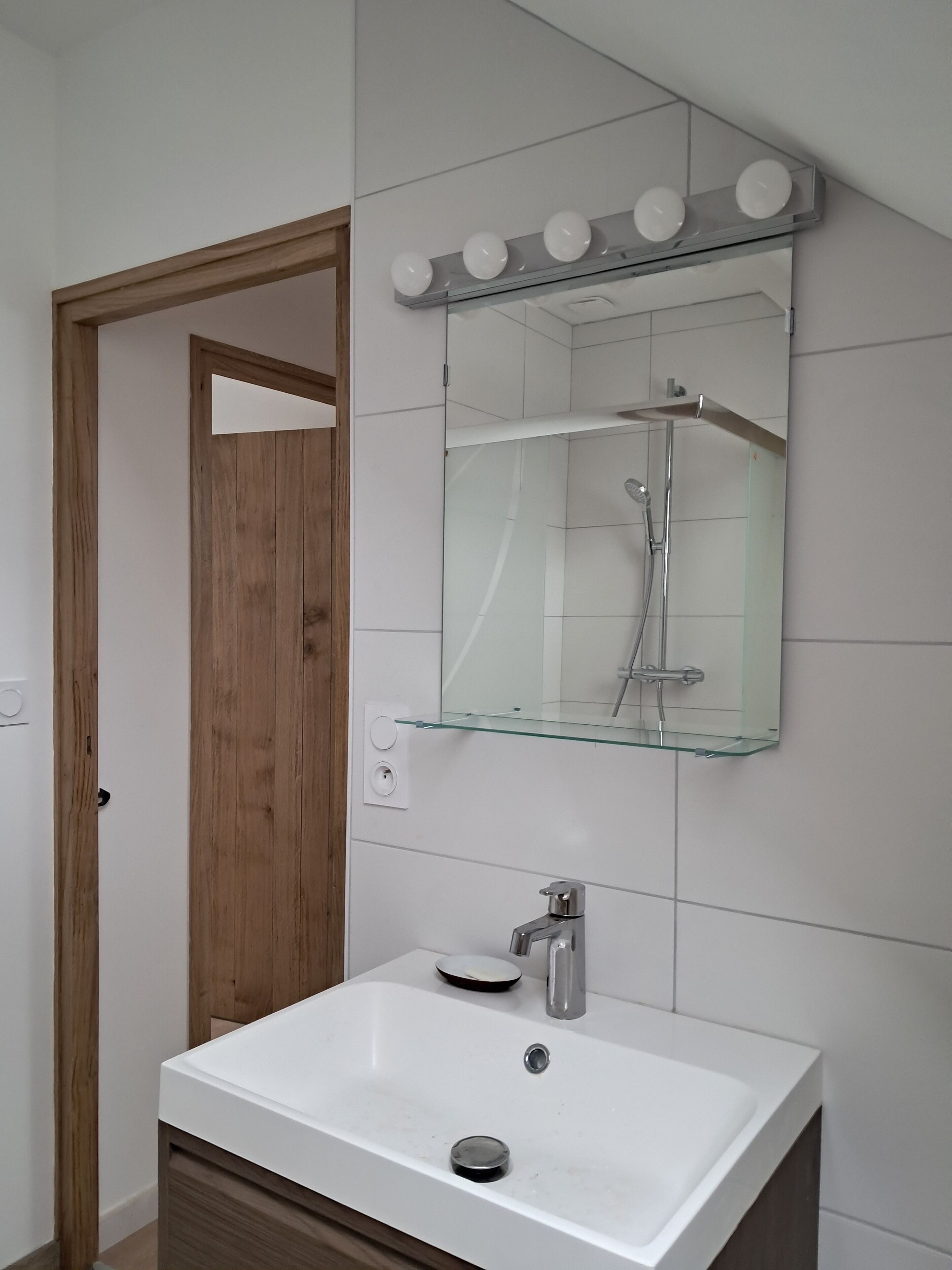 A bathroom with a white sink, a glass shelf above the mirror, and a wooden door.