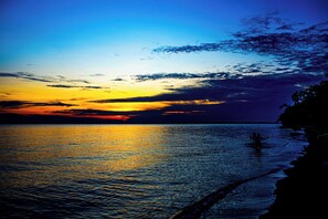 A sunset over a calm sea with a silhouette of a person standing in the water. - Bugoma sand beach resort hotel (Bugala Island)