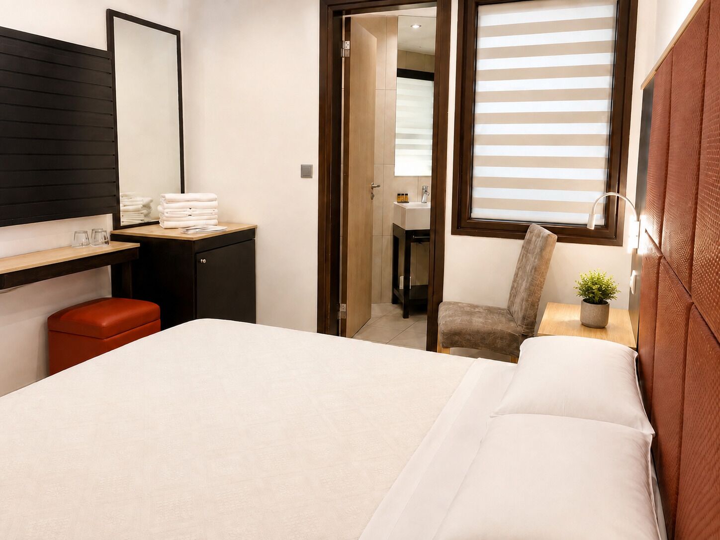 Habitación de hotel con cama, escritorio con espejo, una silla, una mesita con planta y baño visible a través de una puerta abierta.