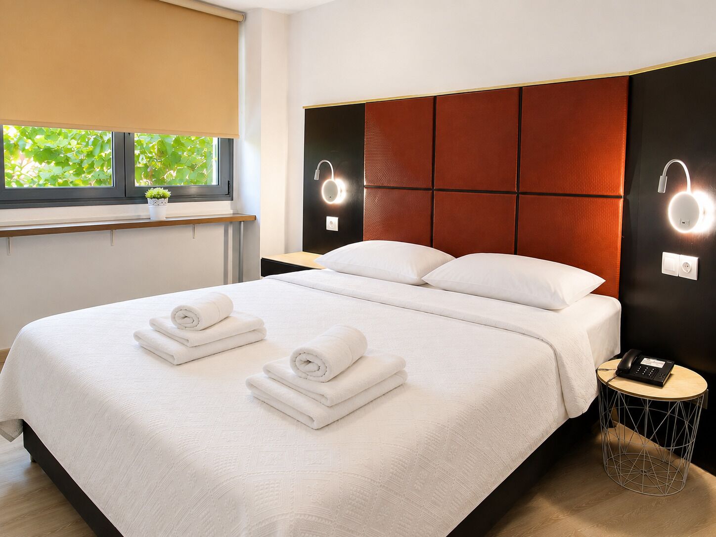 A modern hotel room with a large bed, two bedside tables, a round side table, and a window with a view of greenery.