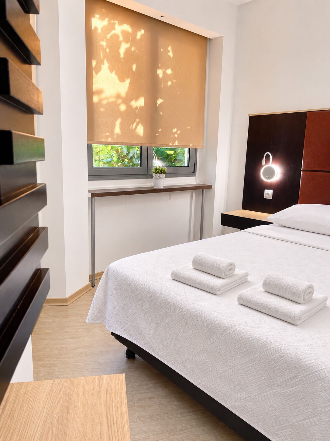 A modern hotel room with a large bed, a window with a view of greenery, a bedside table with a lamp, and a wardrobe.