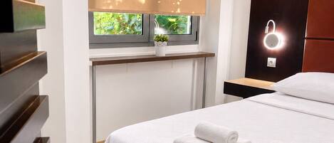 A modern hotel room with a large bed, a window with a view of greenery, a bedside table with a lamp, and a wardrobe.