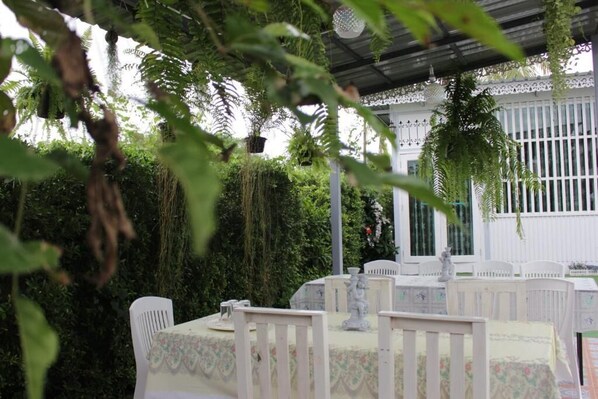 An outdoor dining area with white chairs and tables, surrounded by lush greenery and a white fence. - Moeimanee Resort (Mae Sot)