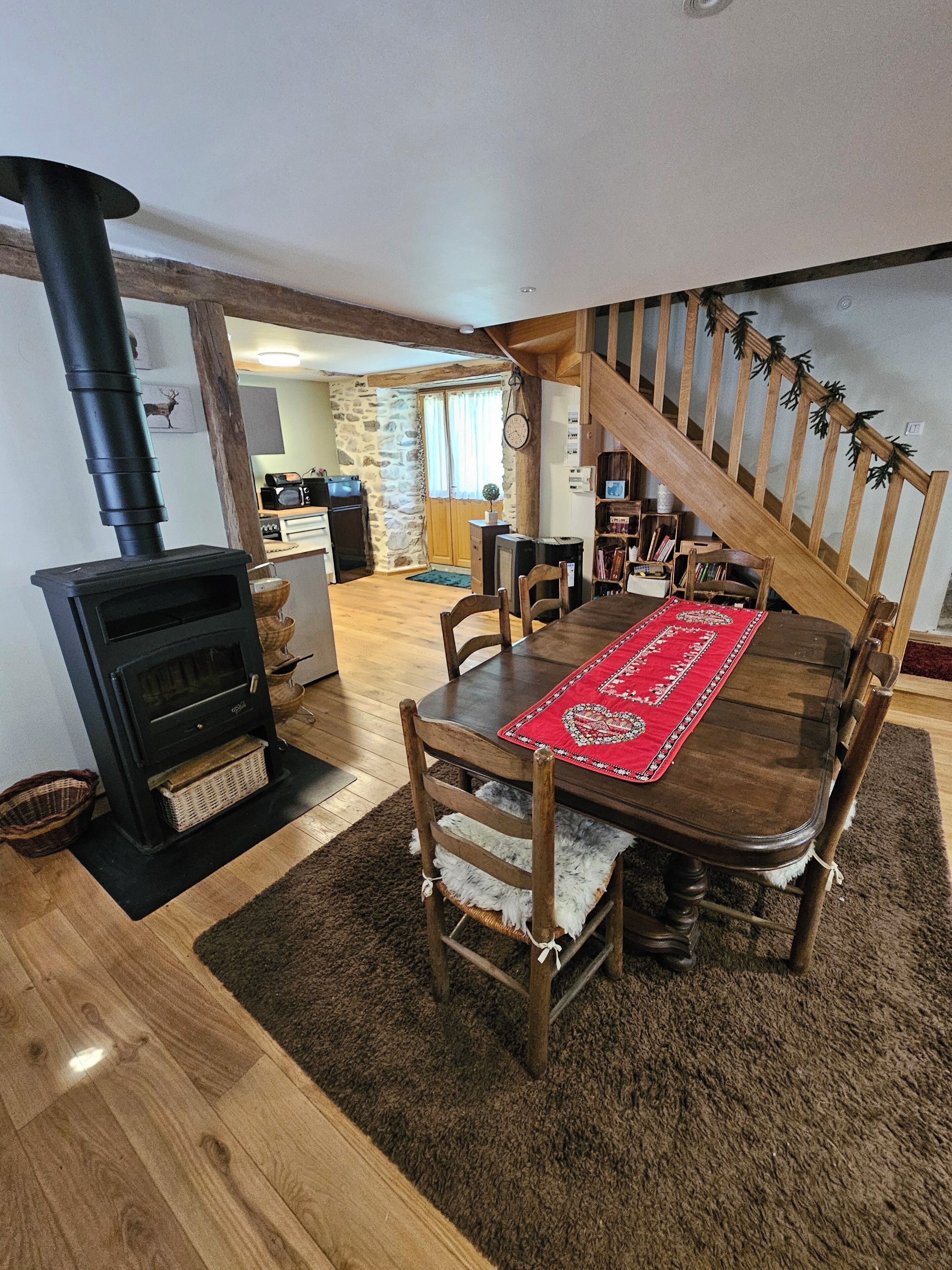 A cozy dining area with a wooden table, chairs, and a red runner. A black wood stove is visible in the foreground.