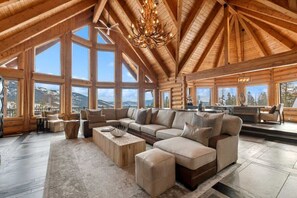 A spacious log cabin interior with large windows, a sectional sofa, and a chandelier.