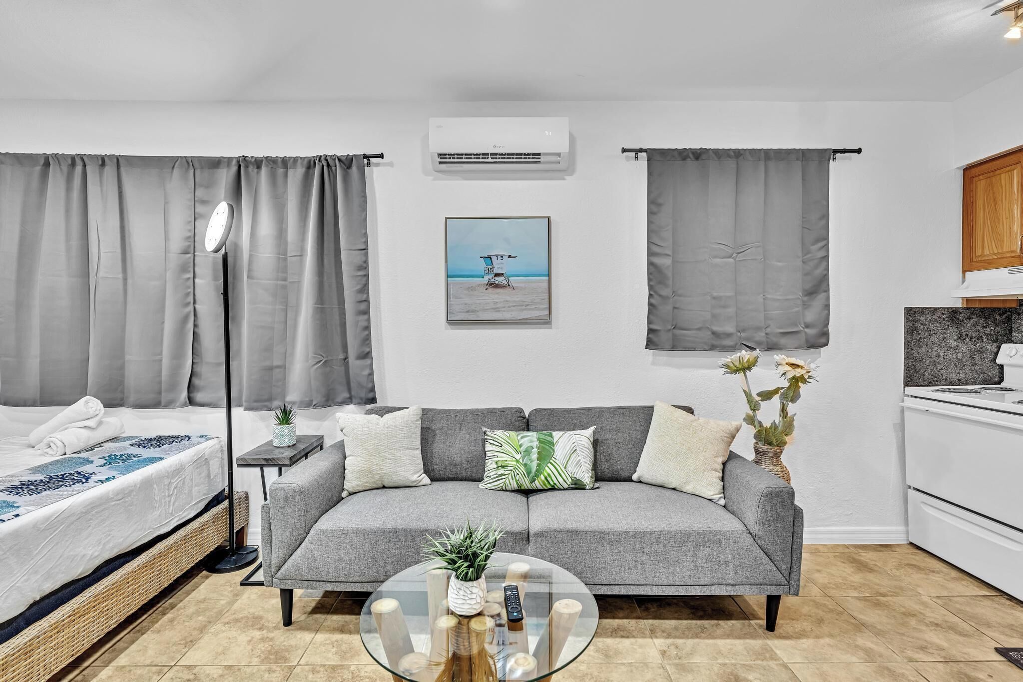 Een moderne, goed verlichte kamer met een grijze bank, een glazen salontafel, een bed met een deken met een wit en blauw patroon, en een lijst met een afbeelding van een strandgezicht.
