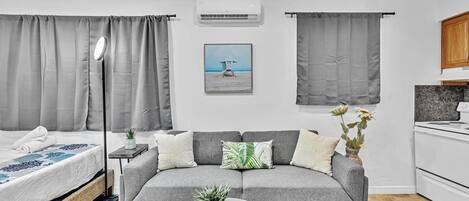 Een moderne, goed verlichte kamer met een grijze bank, een glazen salontafel, een bed met een deken met een wit en blauw patroon, en een lijst met een afbeelding van een strandgezicht.