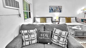 A modern hotel room with a grey sofa, two beds, a painting, and a lamp.