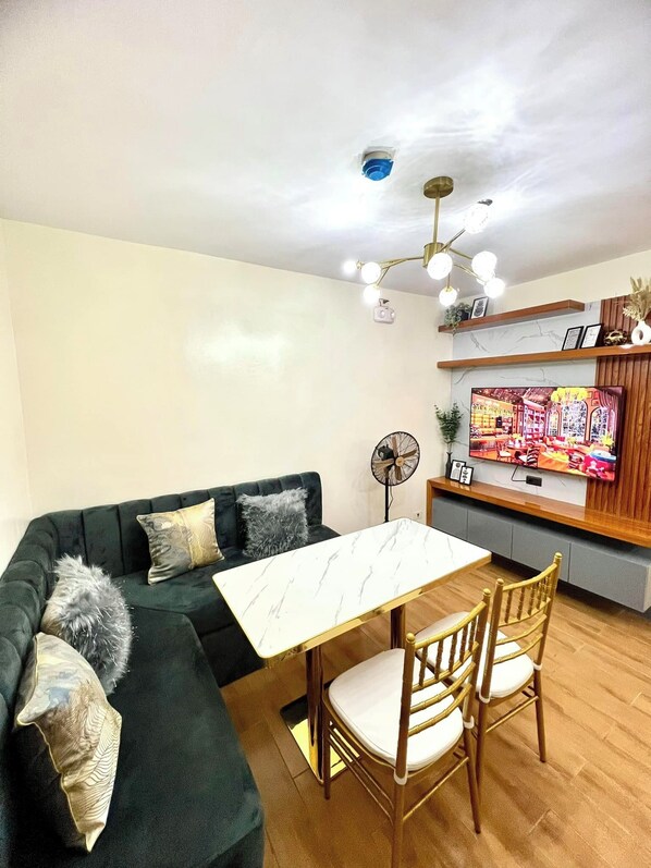 Un salon moderne comprenant un canapé d’angle, une table à manger avec des chaises, un téléviseur fixé au mur et un ventilateur de plafond.