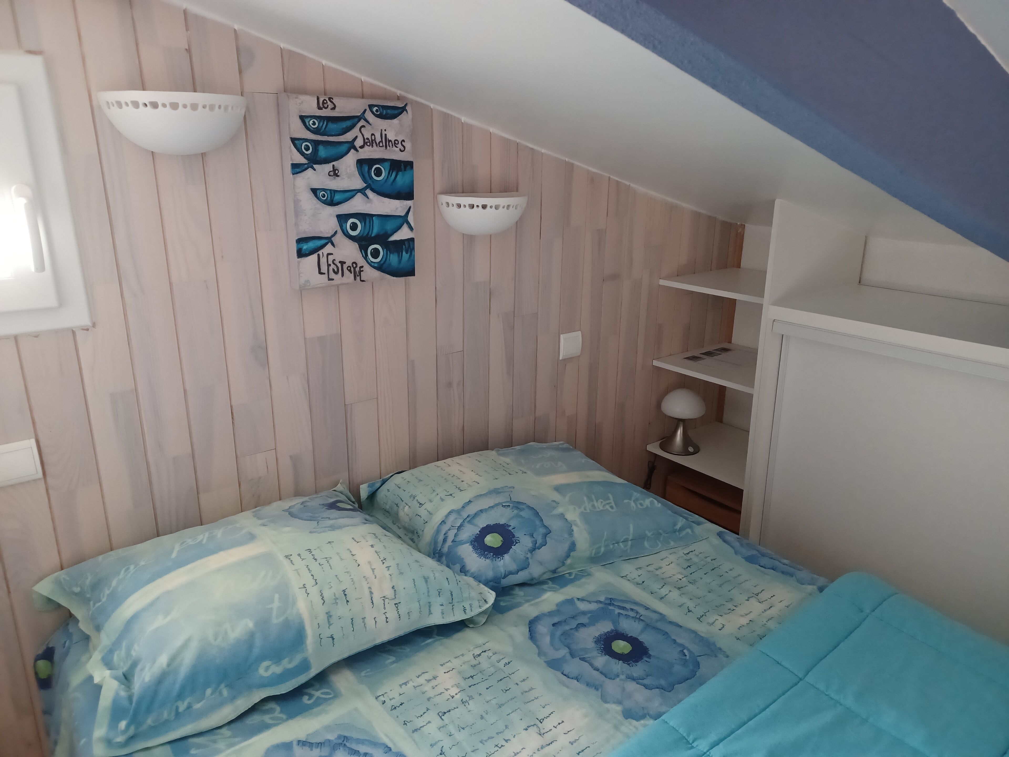 Un dormitorio con techo de listones de madera, una cama con ropa de cama floral azul y repisas fijadas a la pared.