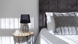 A neatly made bed with a grey quilted headboard and pillows, a bedside table with a lamp and a mug, and a power outlet on the wall.