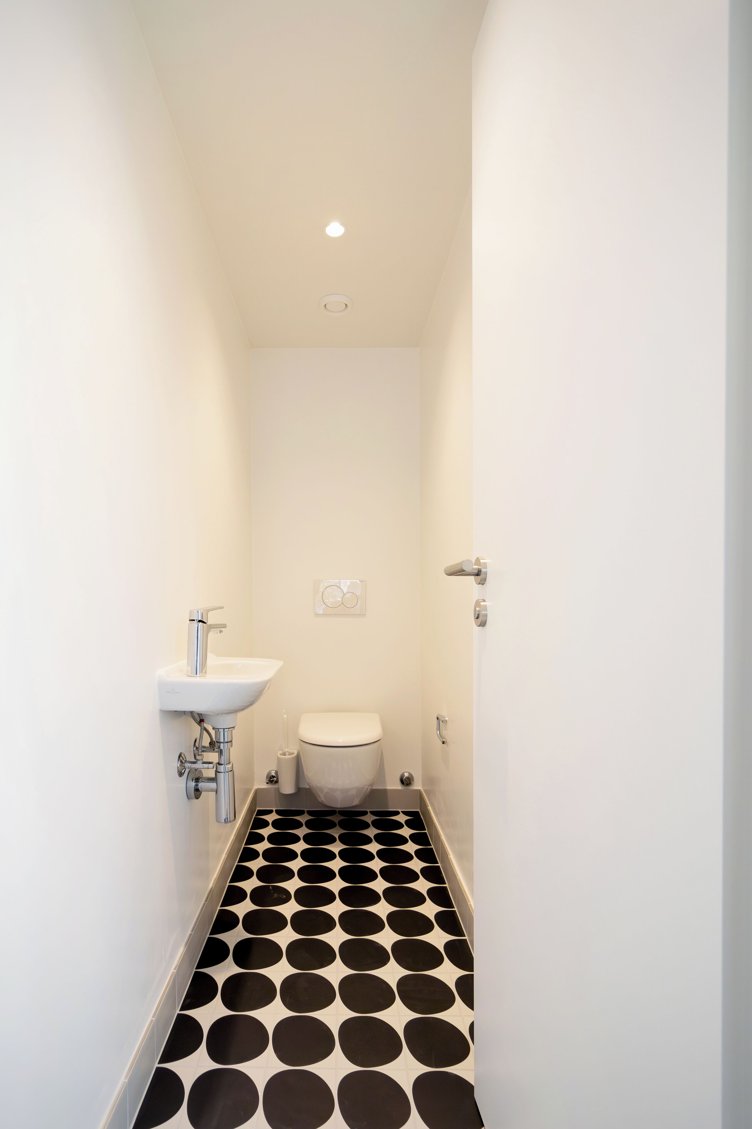 A narrow bathroom with a white sink, toilet, and black and white patterned floor.