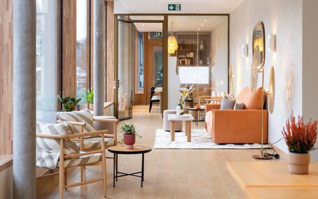 A modern hotel lobby with a wooden floor, a seating area with a sofa and chairs, a small table with a plant, and a large mirror on the wall.