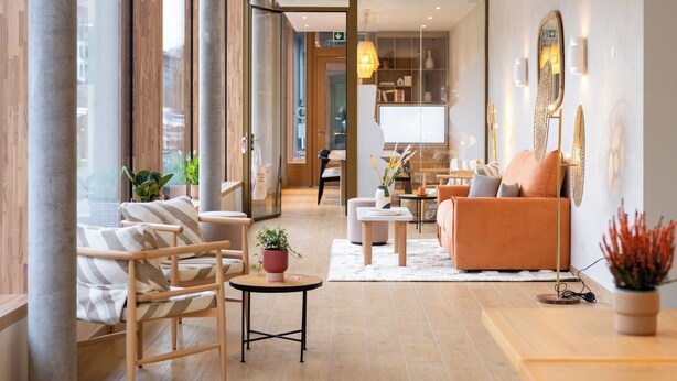 A modern hotel lobby with a wooden floor, a seating area with a sofa and chairs, a small table with a plant, and a large mirror on the wall. - HOOD Hôtel (Geneva)