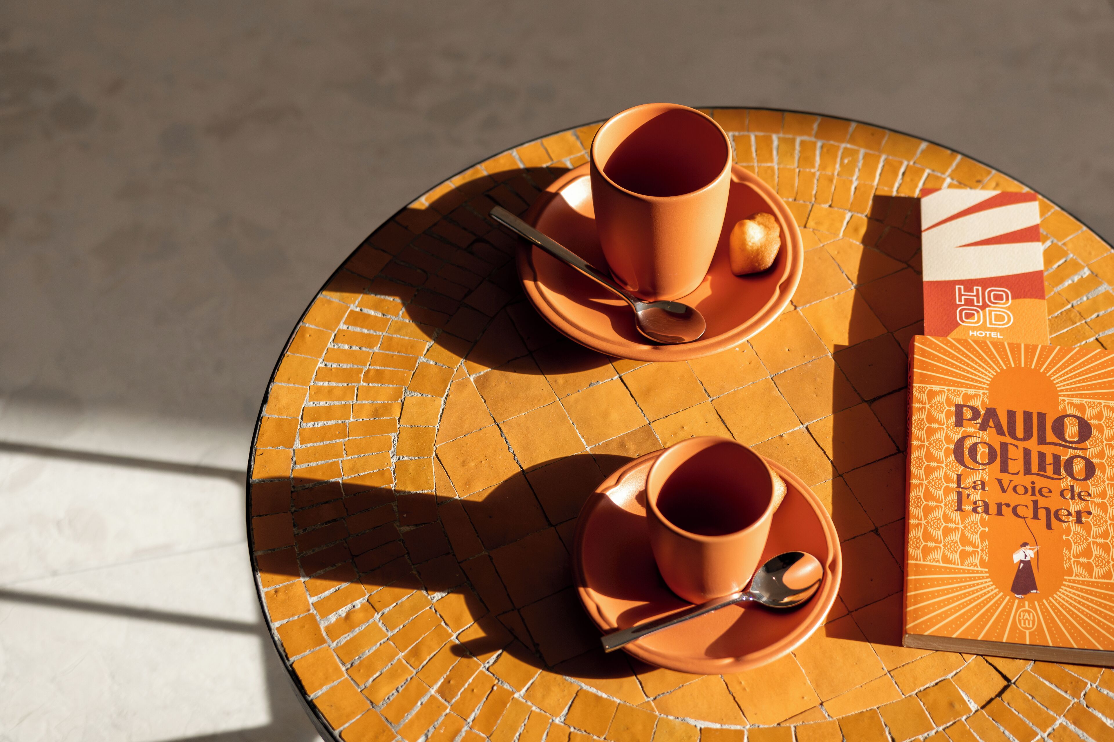 A round table with two matching orange cups and saucers, a spoon, and a book titled "La Voie de l'archer" by Paulo Coelho.