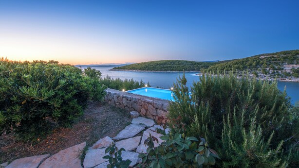 Una piscina rodeada de muros de piedra y exuberante vegetación, con vistas a un cuerpo de agua y colinas distantes.