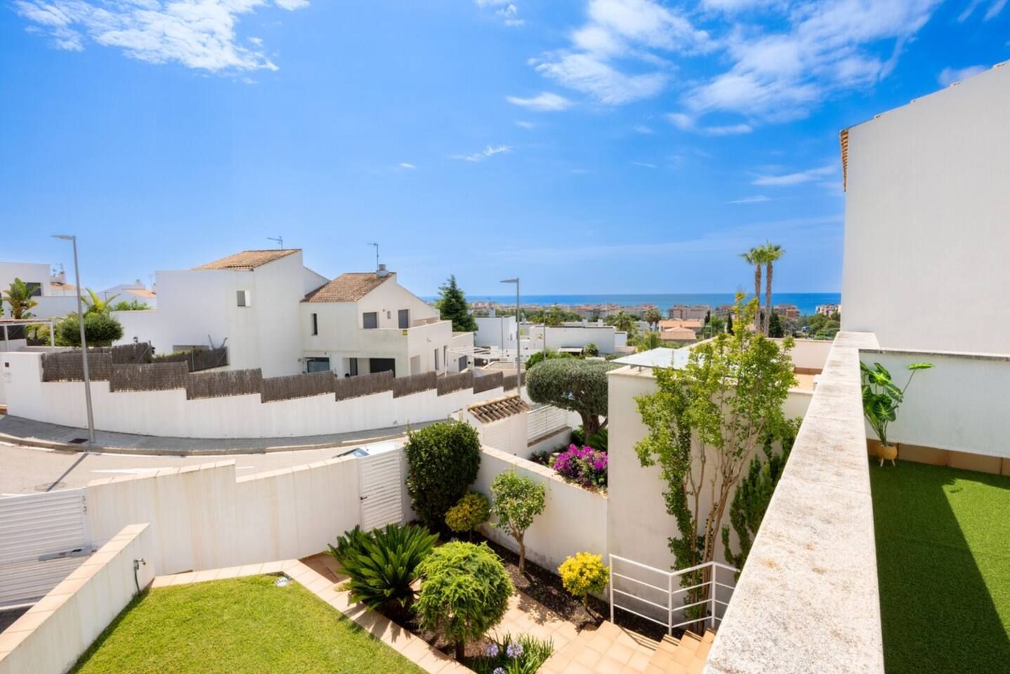 A residential area with white buildings, a small garden with various plants, and a clear view of the sea in the background.