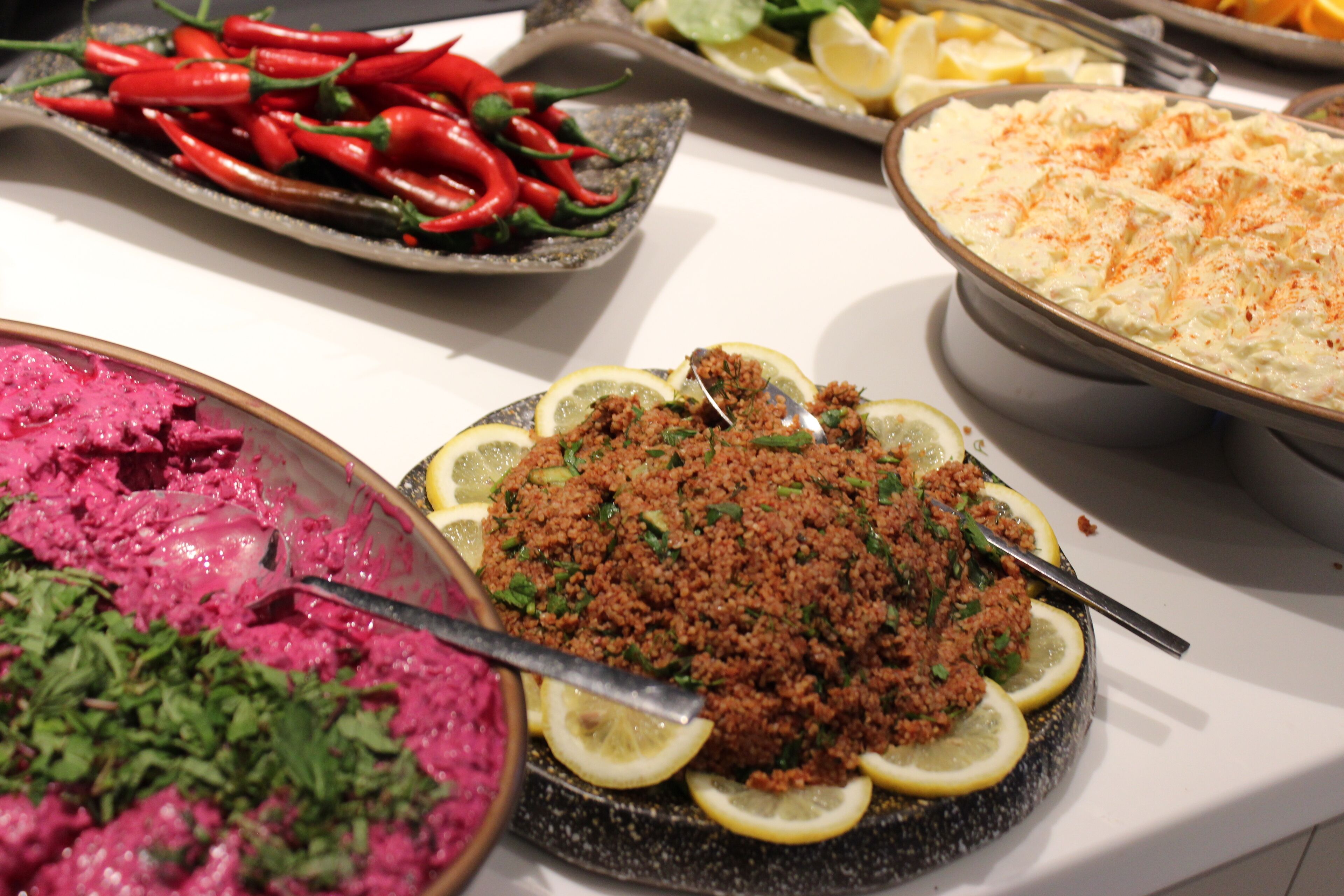 A buffet table with various dishes, including a bowl of pink beet salad, a dish with ground meat garnished with lemon slices, and a large serving of a creamy dish topped with a red spice.