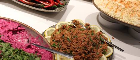 A buffet table with various dishes, including a bowl of pink beet salad, a dish with ground meat garnished with lemon slices, and a large serving of a creamy dish topped with a red spice.