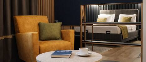 Una camera d'albergo con una grande poltrona, un tavolino con un libro e una tazza, e un letto con cuscini e coperta.