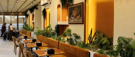 A restaurant with long wooden tables and cushioned booths, surrounded by potted plants and a large painting on the wall.