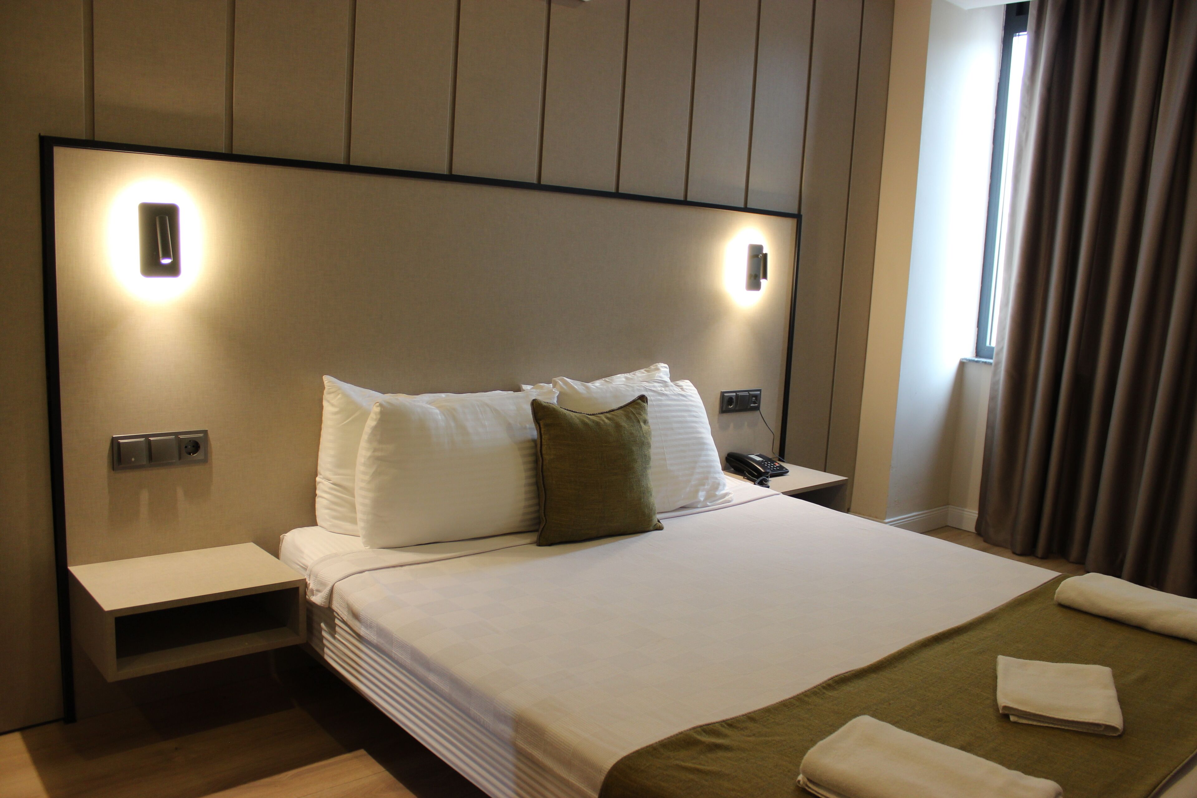 A neatly made hotel room with a large bed, a bedside table, a nightstand, and a wall-mounted light fixture.