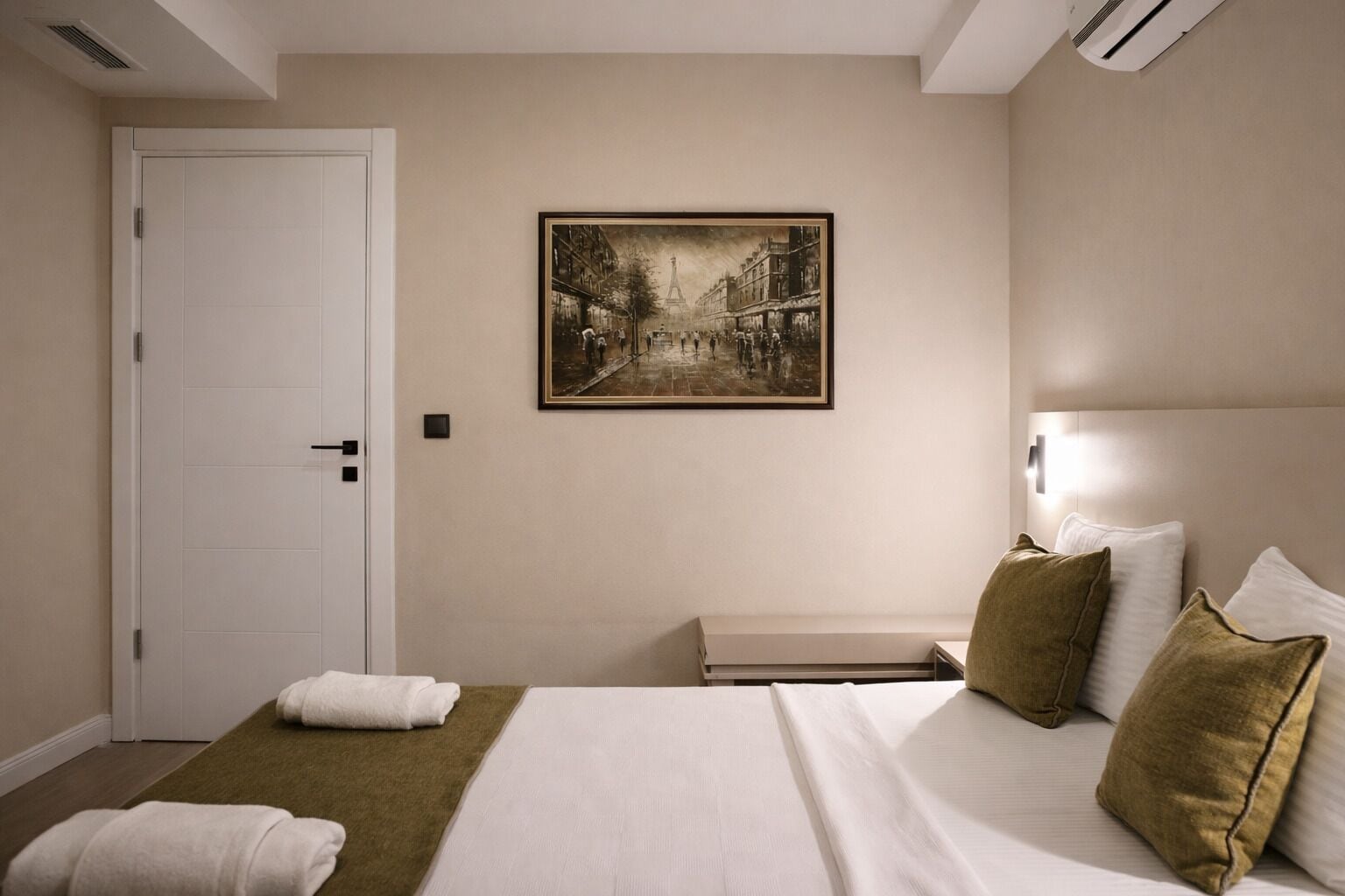 A hotel room with a bed, pillows, a bench, a door, and a framed picture on the wall.