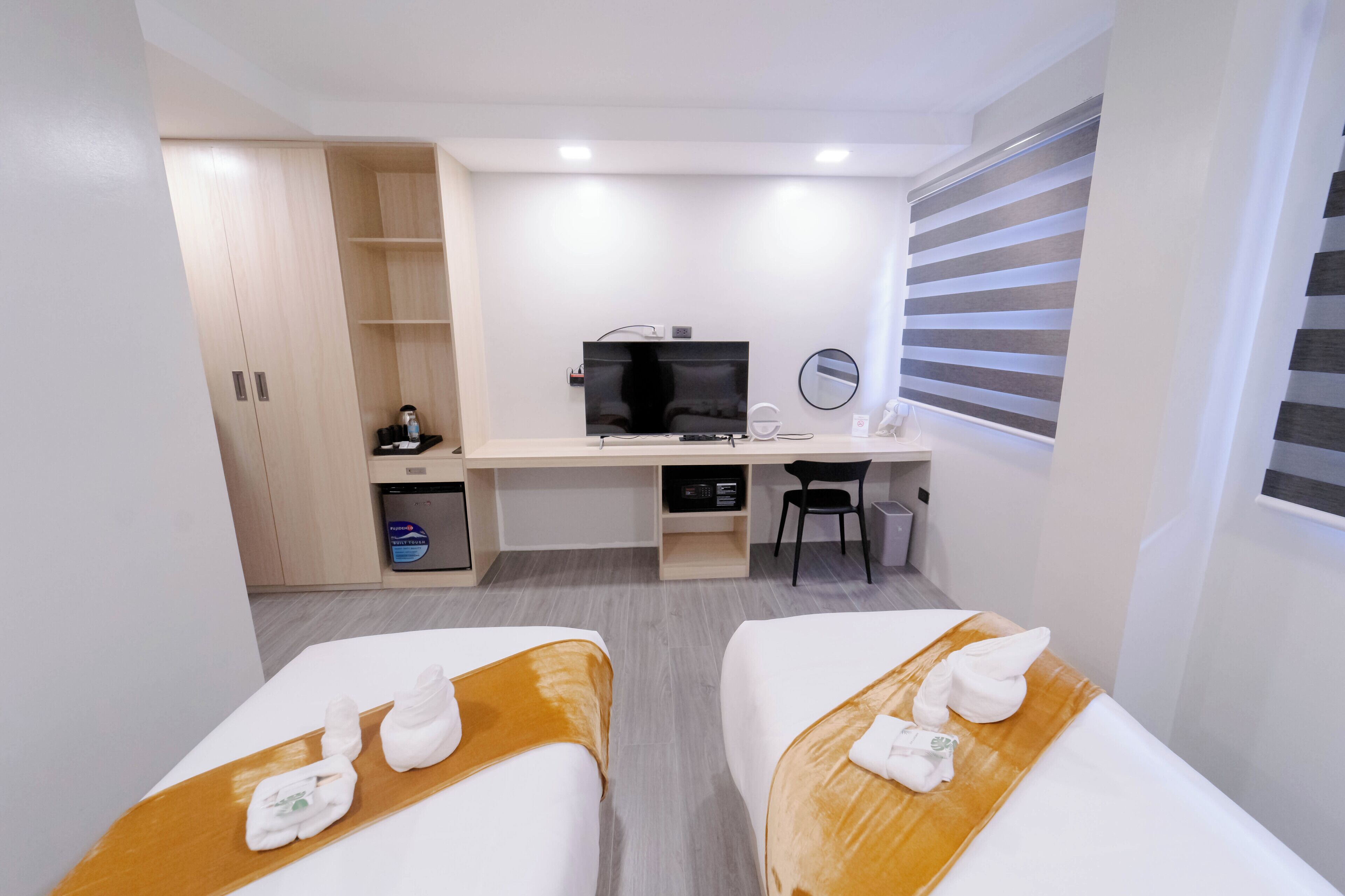 Habitación de hotel con dos camas, un escritorio, un televisor, un espejo y una ventana con persianas.