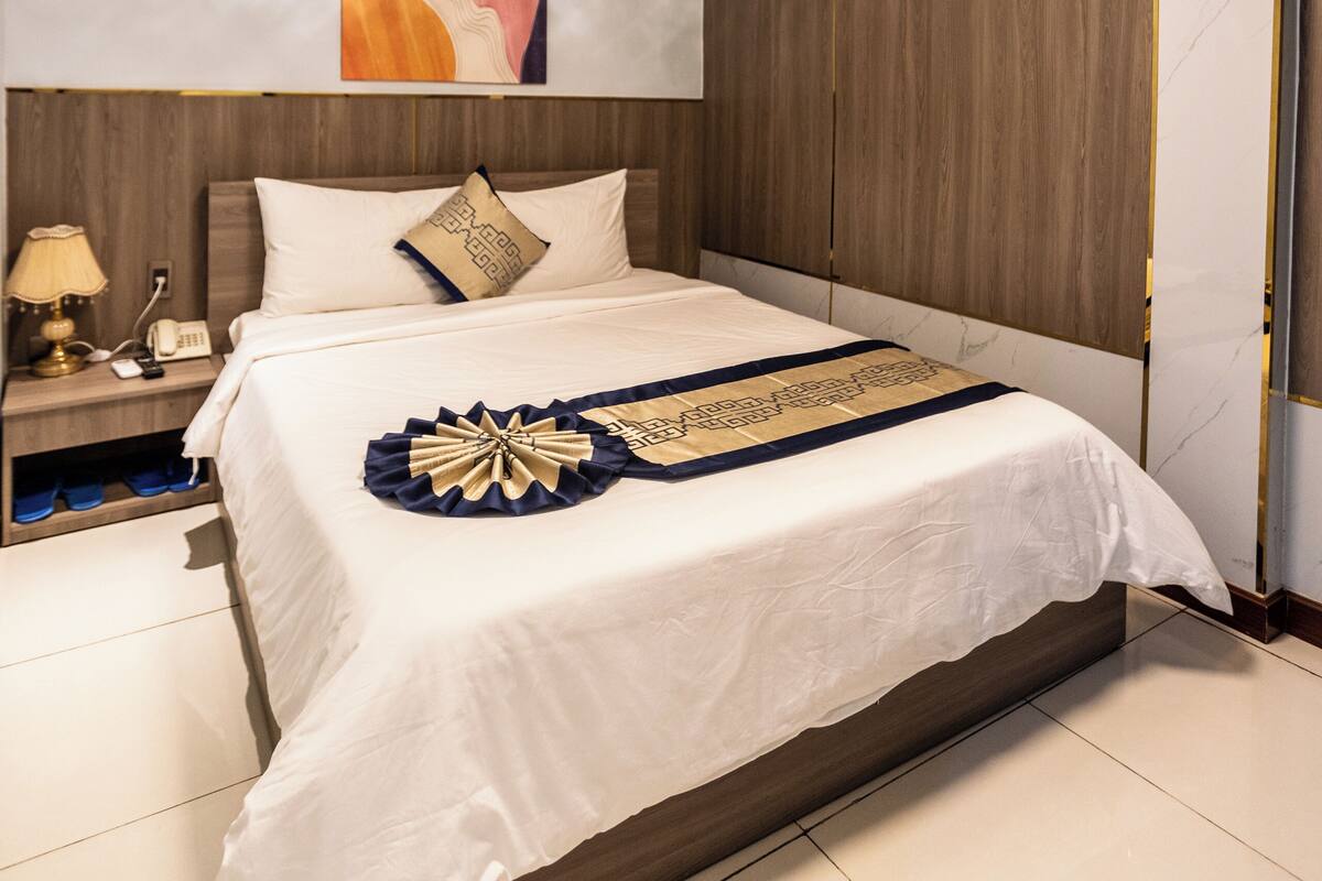 A modern hotel room with a large bed, a wooden headboard, a nightstand with a lamp, and a ceiling light fixture.