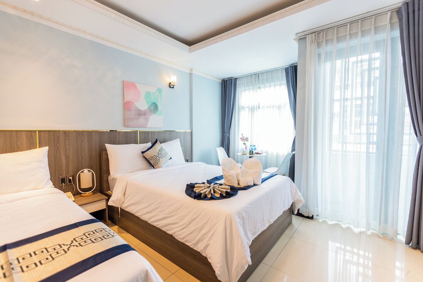 A hotel room with a large bed, a smaller bed, a bedside table with a lamp, a wall-mounted artwork, and a window with curtains.