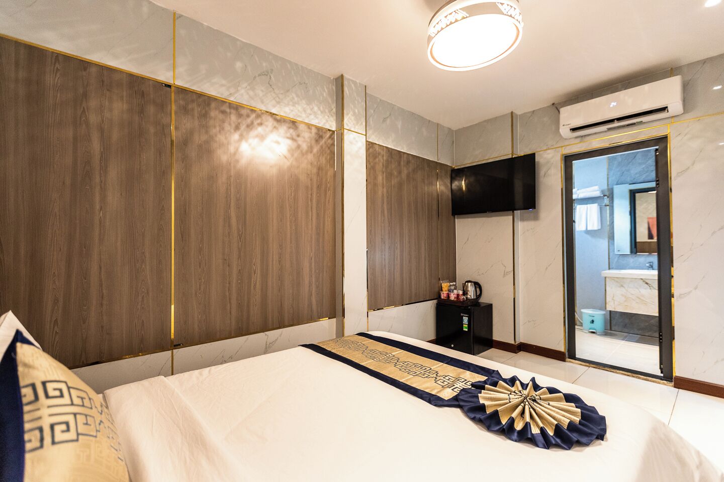 A modern hotel room with a large bed, a flat-screen TV, an air conditioning unit, and a bathroom visible through a glass door.