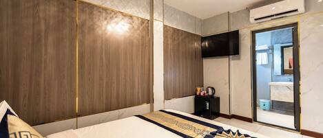 A modern hotel room with a large bed, a flat-screen TV, an air conditioning unit, and a bathroom visible through a glass door.