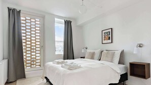 Ein Schlafzimmer mit einem Bett, einem Nachttisch, einer Wandlampe, einem gerahmten Bild und einem Fenster mit Vorhängen.