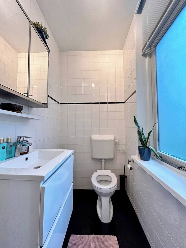 A compact bathroom with white tiles, a toilet, a sink with toiletries, and a window with a plant. - A unique canal-view experience in Amsterdam City Center (Amsterdam)