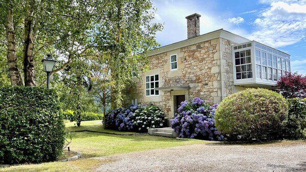 Une maison en pierre dotée d’une grande fenêtre, entourée de haies bien entretenues et de plantes en fleurs.