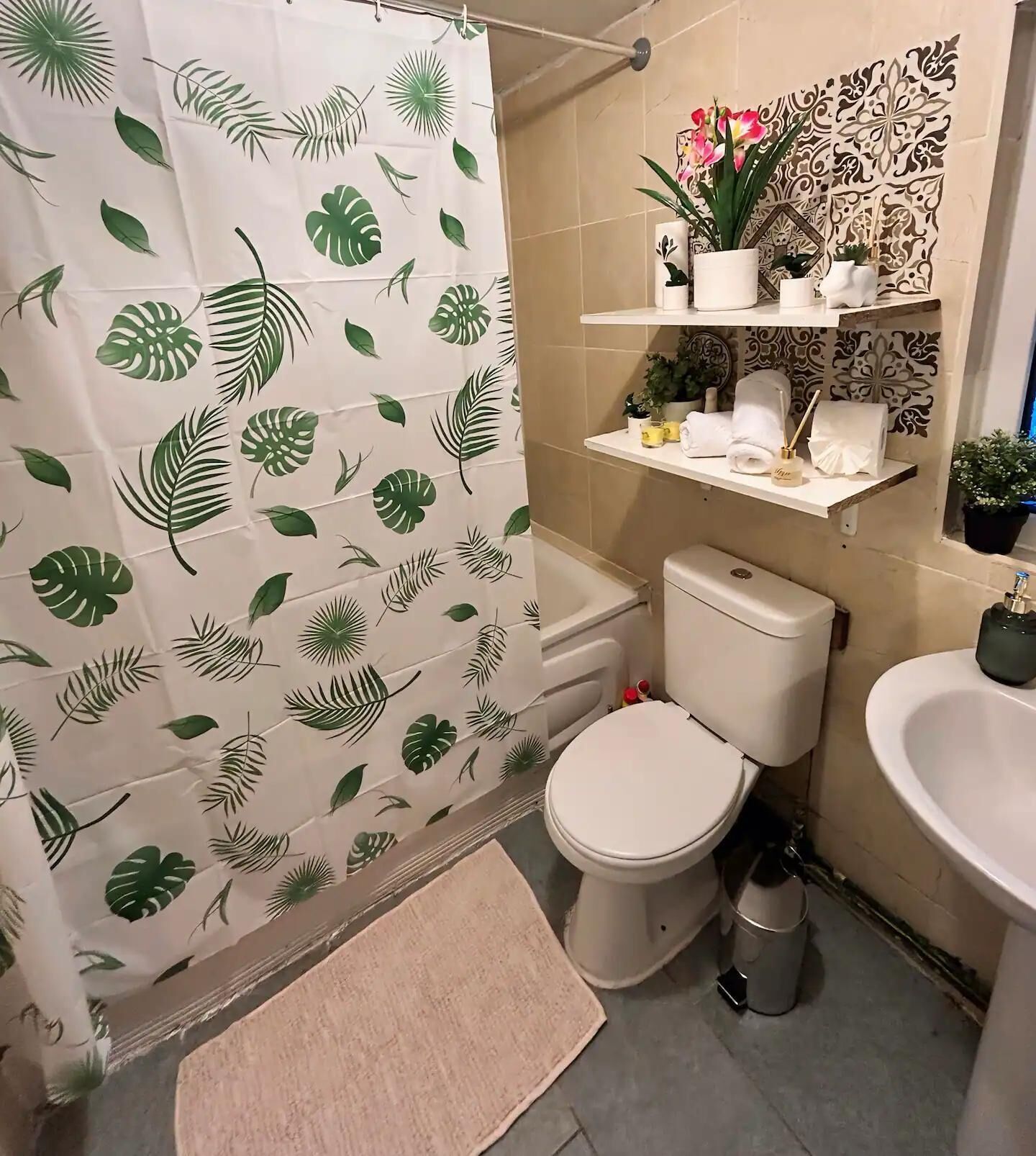 A bathroom with a shower curtain featuring a leaf pattern, a toilet, a sink, and a shelf with toiletries.