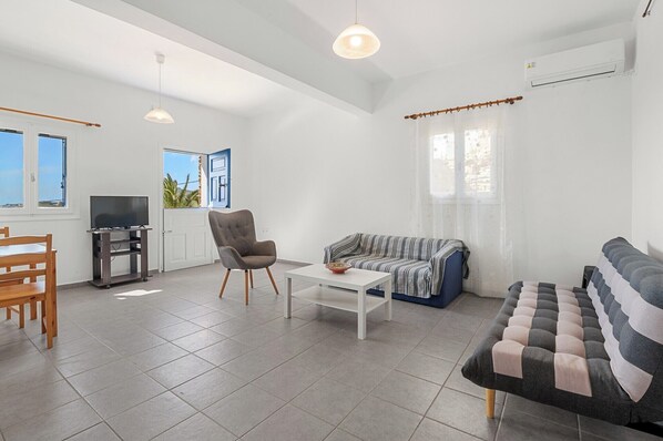 A modern living room with a TV, sofa, armchair, and a dining area with a wooden table and chairs. - Family House for 3 by Tinosstay (Tinos)