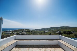 Un balcon doté d’un sol en pierre, offrant une vue sur un village côtier avec ses maisons et un ciel dégagé.