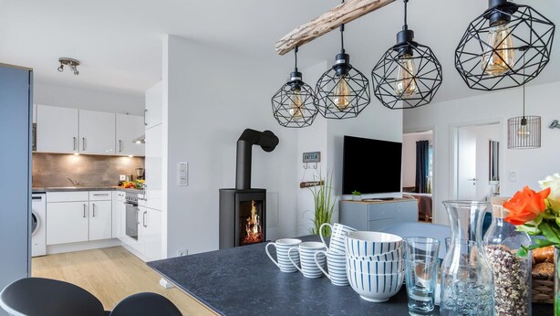 A modern kitchen with white cabinets, a black countertop, and a central island with seating. There is a fireplace, a TV mounted on the wall, and a hanging light fixture above the island. - Beautiful home in Olpenitz with sauna (Olpenitz)